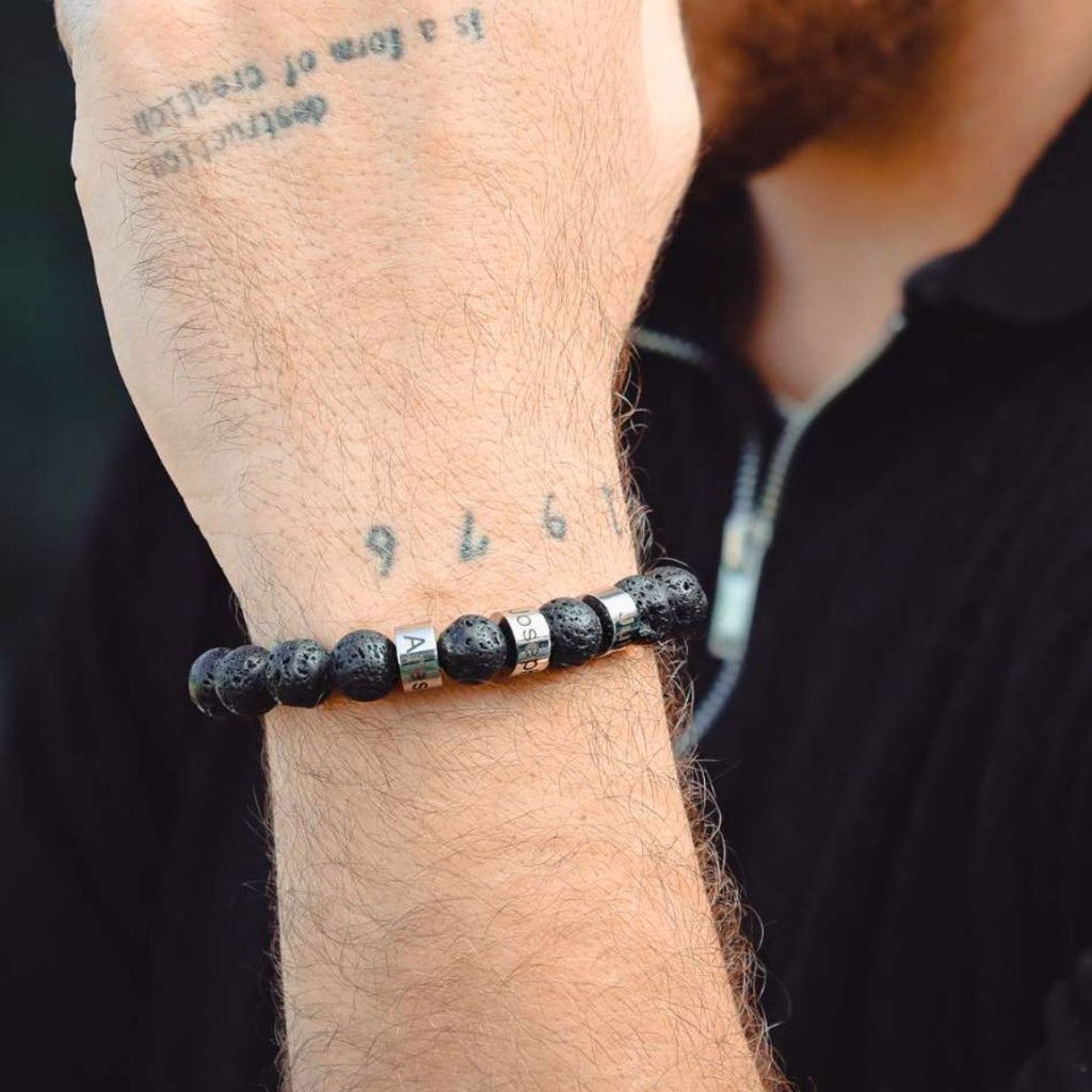Un primer plano de una muñeca con un tatuaje 976 lleva una Pulsera de piedra volcánica personalizada con cuentas volcánicas negras y cuentas de letras plateadas, combinada con un top oscuro con cremallera. Ideal como pulsera personalizada para papá.