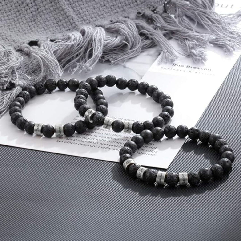 Tres pulseras de piedra volcánica personalizadas con cuentas plateadas se exhiben sobre una superficie con papeles y tela gris claro al fondo, ideales como regalo día del padre o pulsera personalizada para papá.