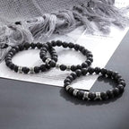 Tres pulseras de piedra volcánica personalizadas con cuentas plateadas se exhiben sobre una superficie con papeles y tela gris claro al fondo, ideales como regalo día del padre o pulsera personalizada para papá.
