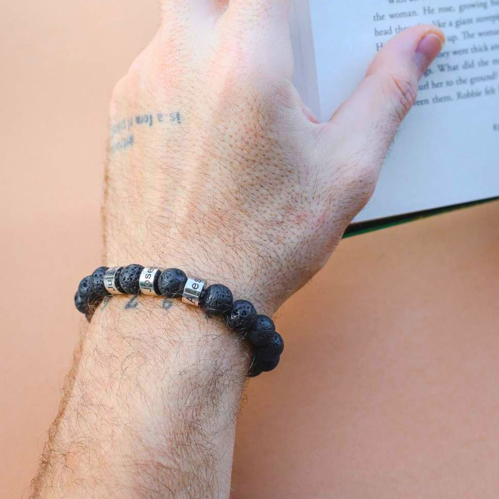 Una mano tatuada lleva una Pulsera de piedra volcánica personalizada con cuentas de letras plateadas que deletrean BEST SIS mientras sostiene un libro abierto sobre un fondo melocotón.
