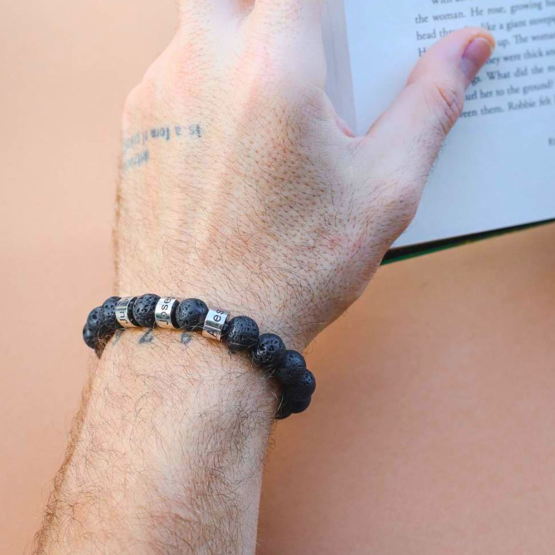 Una mano tatuada lleva una Pulsera de piedra volcánica personalizada con cuentas de letras plateadas que deletrean BEST SIS mientras sostiene un libro abierto sobre un fondo melocotón.
