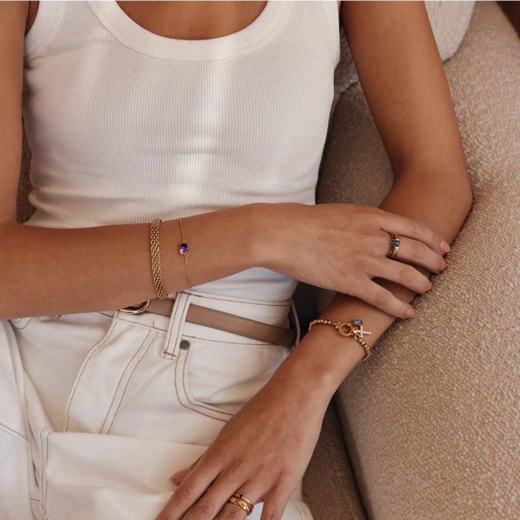 Una persona con camiseta blanca de tirantes y vaqueros está sentada en un sofá beige, con los brazos cruzados, lleva pulseras de oro -incluida la Pulsera dos mitades con piedras de nacimiento-, un anillo de oro, un cinturón de cadena y accesorios con piedras preciosas moradas.