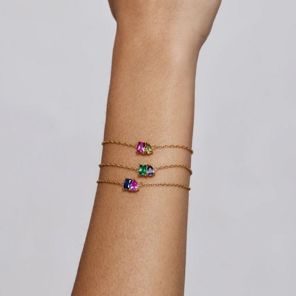 Primer plano de un antebrazo con la Pulsera dos mitades con piedras de nacimiento: una delicada pulsera de cadena de oro con coloridas piedras de nacimiento -ideal como pulsera de amor o joya personalizada- sobre un fondo claro liso.