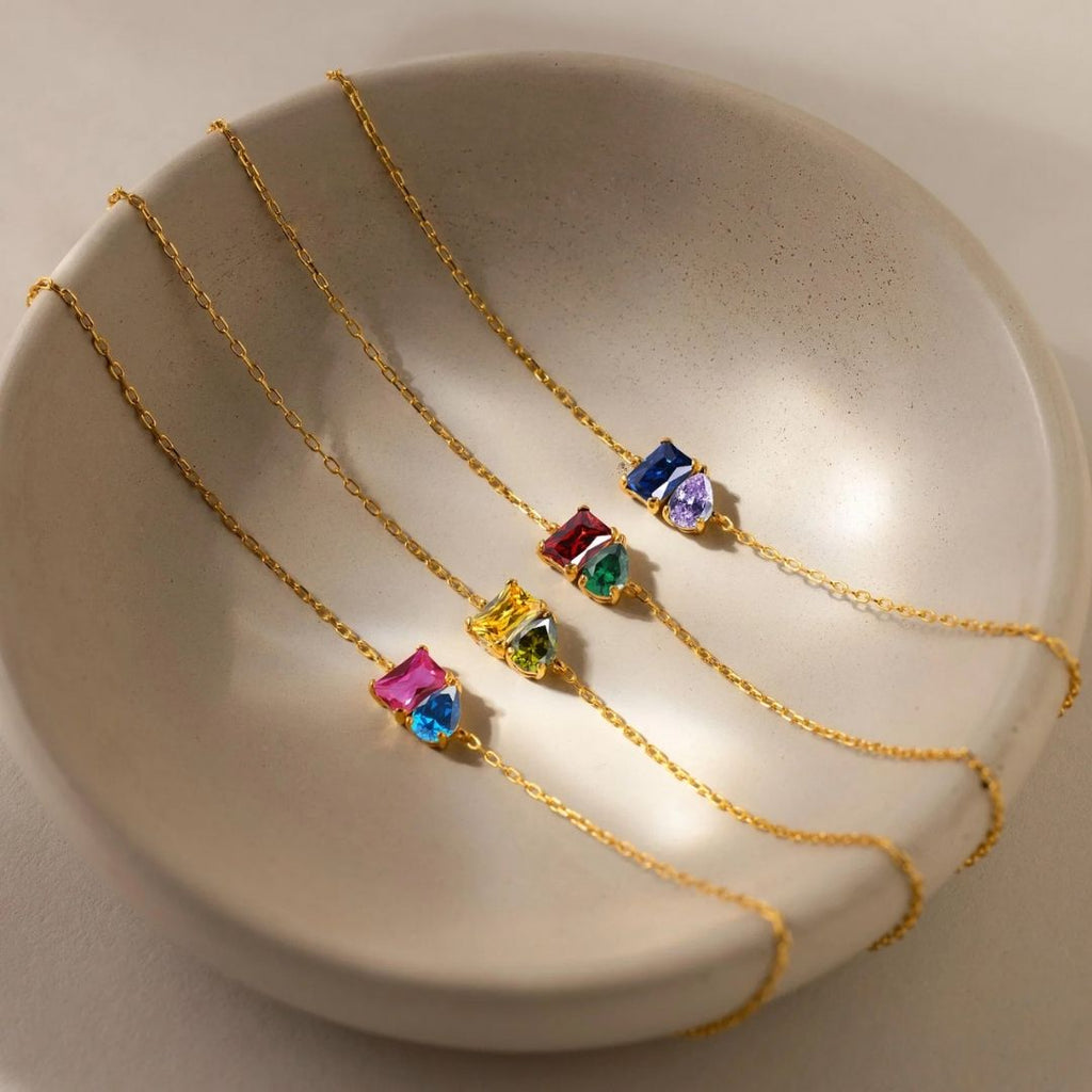 Cuatro delicadas Pulsera dos mitades de oro con piedras de nacimiento, cada una con vibrantes dijes de piedras preciosas en rosa, azul, verde, amarillo y morado, se exhiben en un plato de cerámica beige, perfecto como joya personalizada o pulsera de amor.