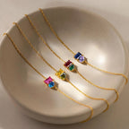 Cuatro delicadas Pulsera dos mitades de oro con piedras de nacimiento, cada una con vibrantes dijes de piedras preciosas en rosa, azul, verde, amarillo y morado, se exhiben en un plato de cerámica beige, perfecto como joya personalizada o pulsera de amor.