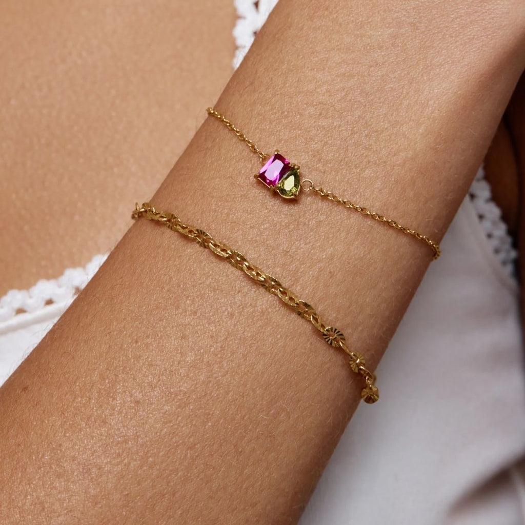Primer plano del brazo de una persona que lleva dos pulseras de oro -una Pulsera dos mitades con piedras de nacimiento y una delicada cadena trenzada- combinadas con un top blanco ribeteado de encaje.
