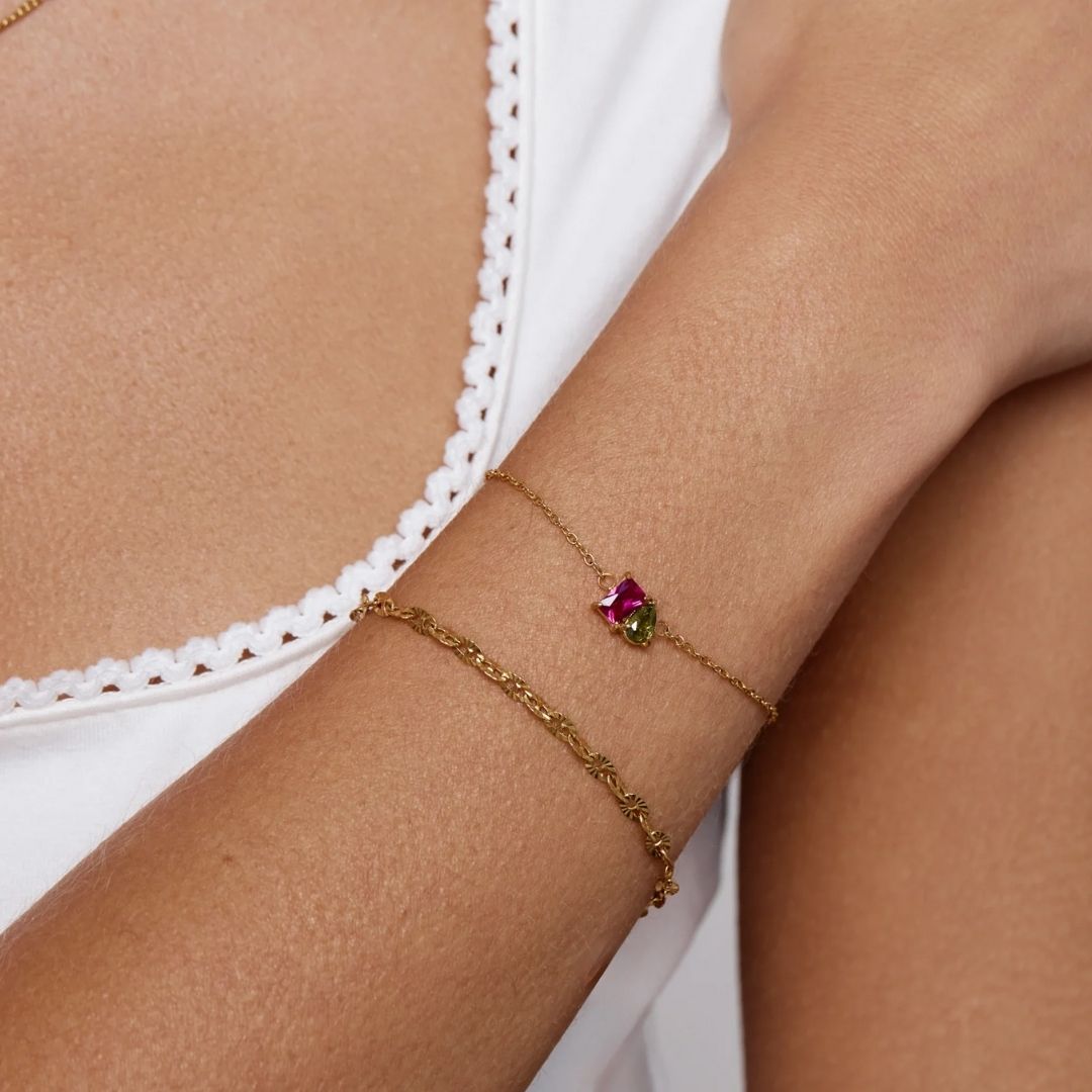 Una persona con un top blanco de cuello festoneado lleva dos pulseras de oro: una sencilla cadena texturizada y la Pulsera dos mitades con piedras de nacimiento con gemas rosas y verdes.