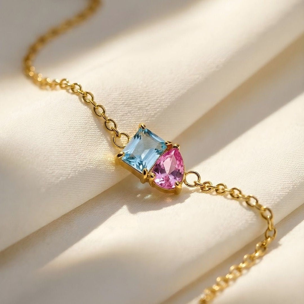 La Pulsera dos mitades con piedras de nacimiento presenta dos mitades de piedras de nacimiento en una delicada cadena dorada, que descansan sobre un suave tejido crema, un complemento perfecto para tu colección de joya personalizada.