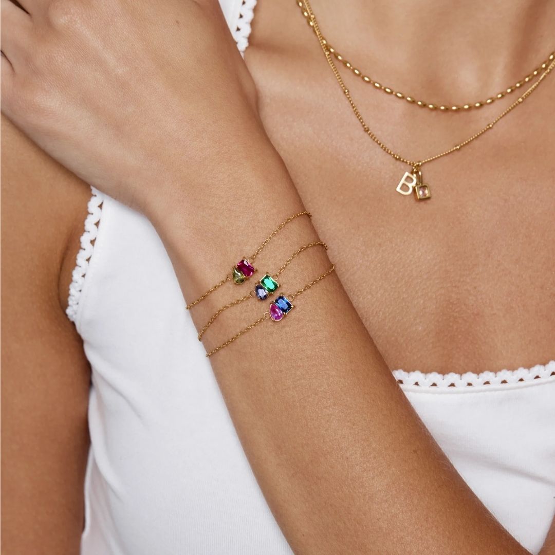 Una mujer con camiseta blanca de tirantes muestra tres pulseras de oro con colgantes de piedras preciosas de colores, incluida la Pulsera dos mitades con piedras de nacimiento, y collares de oro, uno de ellos con un colgante personalizado de la letra B.