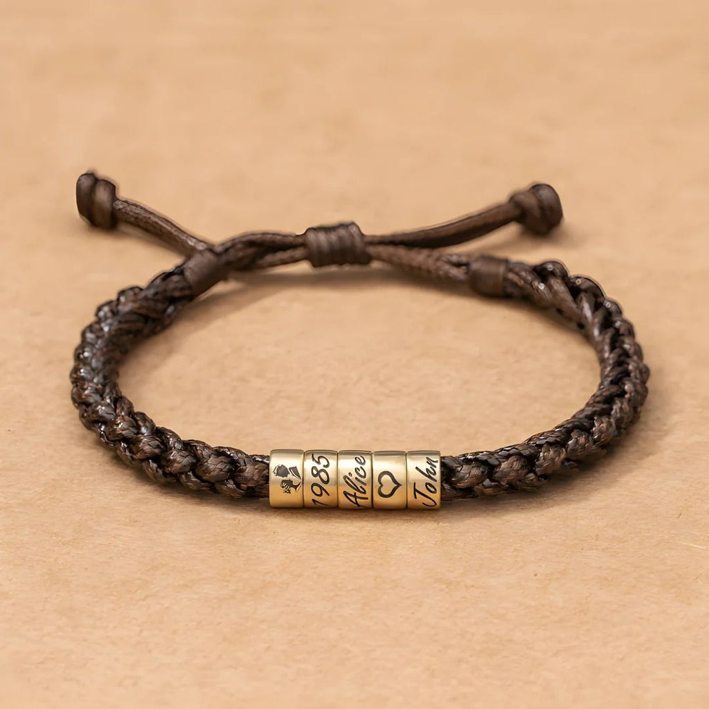 La Pulsera "El mejor Papá" - Personalizable es una pulsera marrón trenzada con tres cuentas doradas grabadas con un trébol de cuatro hojas, 1985, Alicia, un símbolo de corazón y Juan, ideal como pulsera personalizada papá o regalo día del padre.