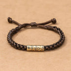 La Pulsera "El mejor Papá" - Personalizable es una pulsera marrón trenzada con tres cuentas doradas grabadas con un trébol de cuatro hojas, 1985, Alicia, un símbolo de corazón y Juan, ideal como pulsera personalizada papá o regalo día del padre.