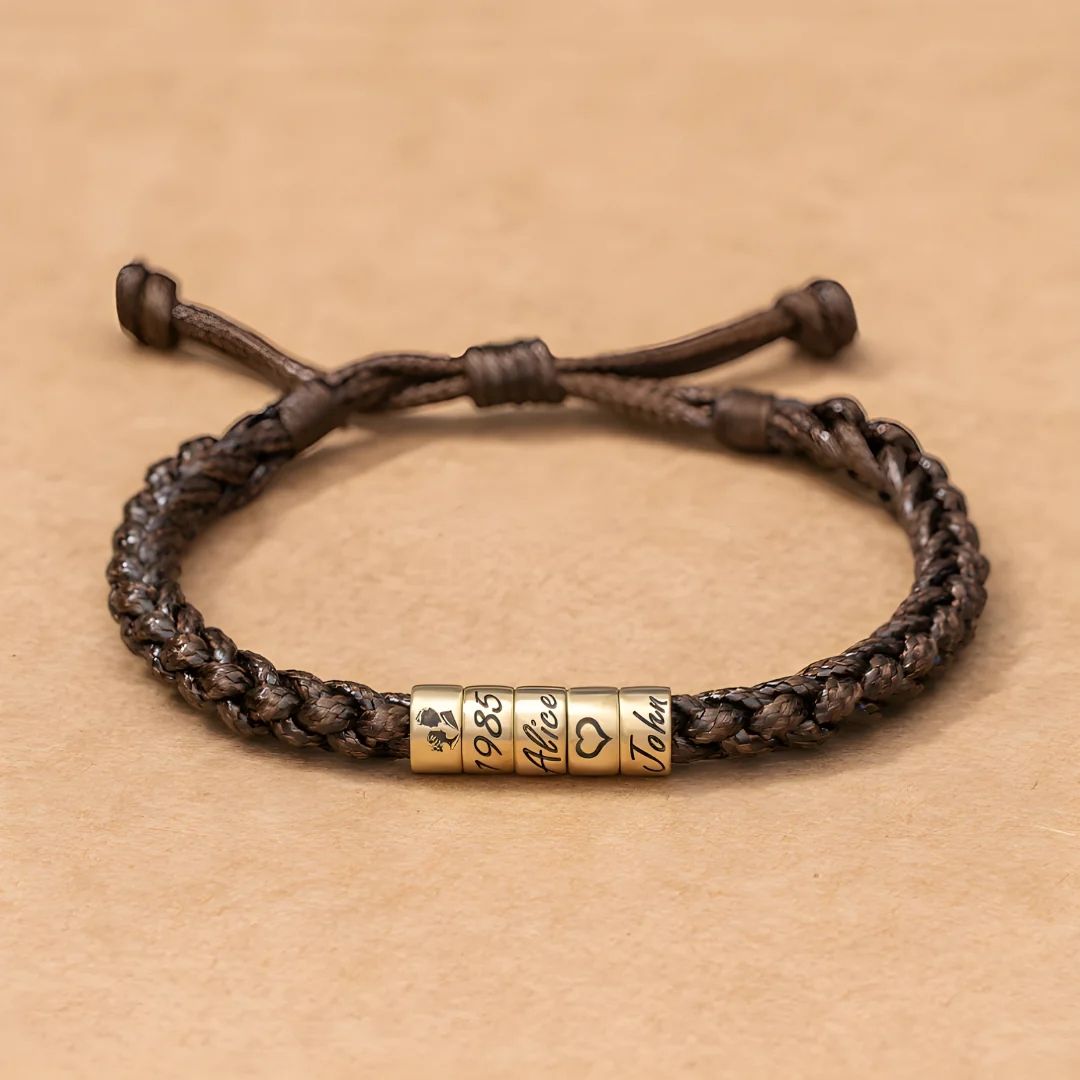 La Pulsera "El mejor Papá" - Personalizable es una pulsera marrón trenzada con tres cuentas doradas grabadas con un trébol de cuatro hojas, 1985, Alicia, un símbolo de corazón y Juan, ideal como pulsera personalizada papá o regalo día del padre.