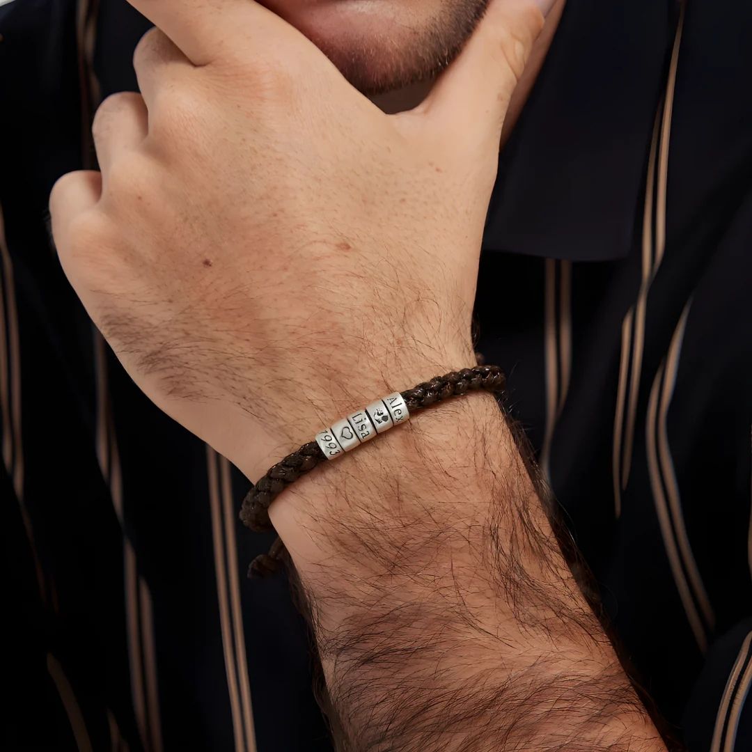 Una persona con camisa de rayas oscuras apoya la barbilla en la mano, mostrando la Pulsera "El mejor Papá" - Personalizable, una pulsera trenzada marrón con cuentas plateadas grabadas, perfecta como regalo el día del padre.