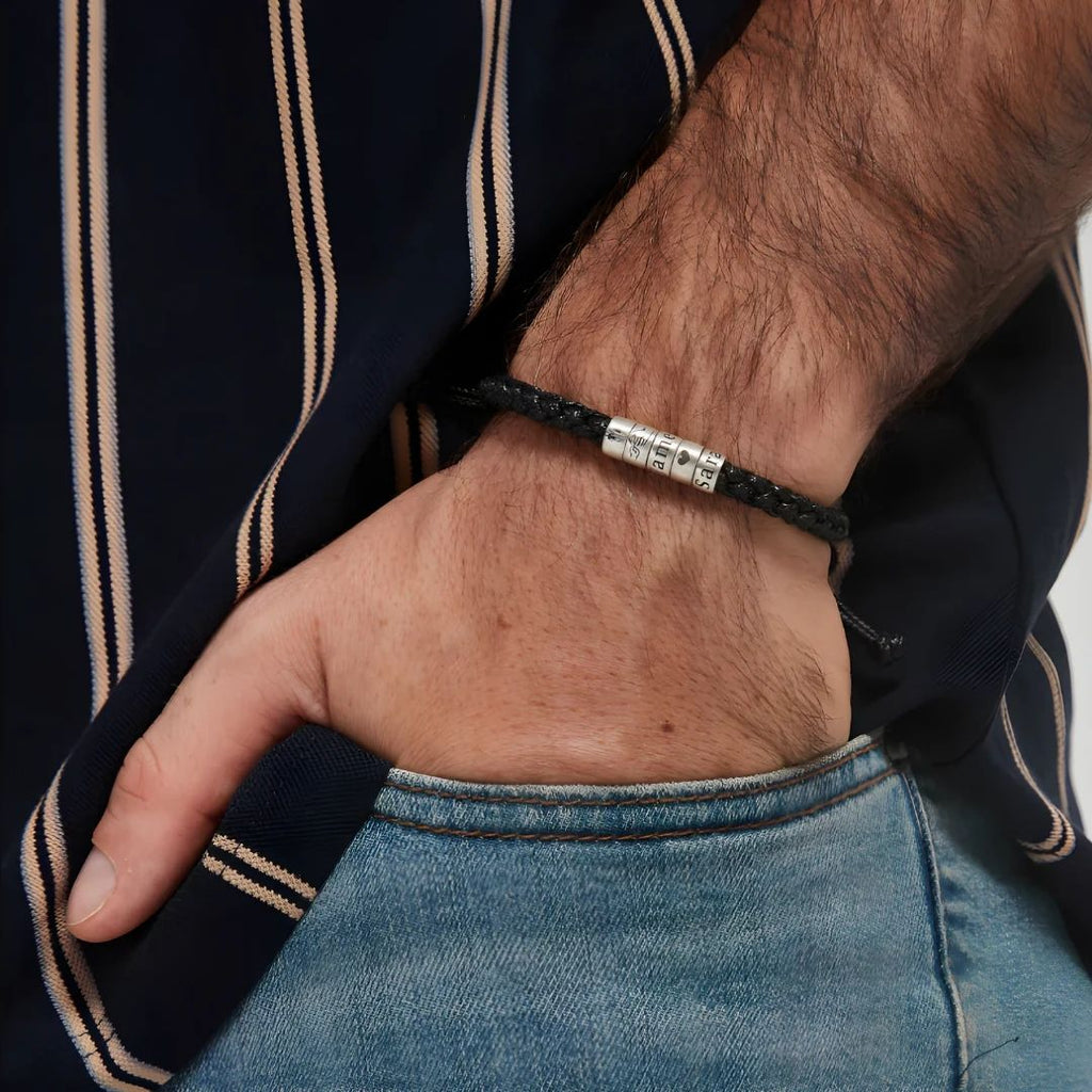 Un primer plano de una mano en el bolsillo de unos vaqueros muestra la Pulsera "El mejor Papá" - Personalizable, una pulsera trenzada negra con detalles plateados, que se lleva con una camisa oscura a rayas-perfecta como regalo día del padre.