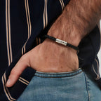 Un primer plano de una mano en el bolsillo de unos vaqueros muestra la Pulsera "El mejor Papá" - Personalizable, una pulsera trenzada negra con detalles plateados, que se lleva con una camisa oscura a rayas-perfecta como regalo día del padre.