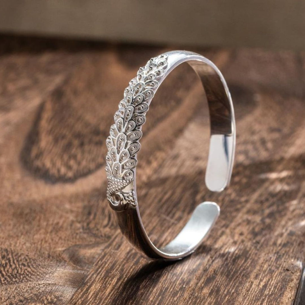 La Pulsera Elegancia Real es una pulsera de plata con un intrincado diseño de plumas de pavo real en un lado, elegantemente expuestas sobre una superficie de madera.