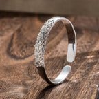 La Pulsera Elegancia Real es una pulsera de plata con un intrincado diseño de plumas de pavo real en un lado, elegantemente expuestas sobre una superficie de madera.