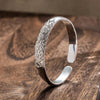 La Pulsera Elegancia Real es una pulsera de plata con un intrincado diseño de plumas de pavo real en un lado, elegantemente expuestas sobre una superficie de madera.