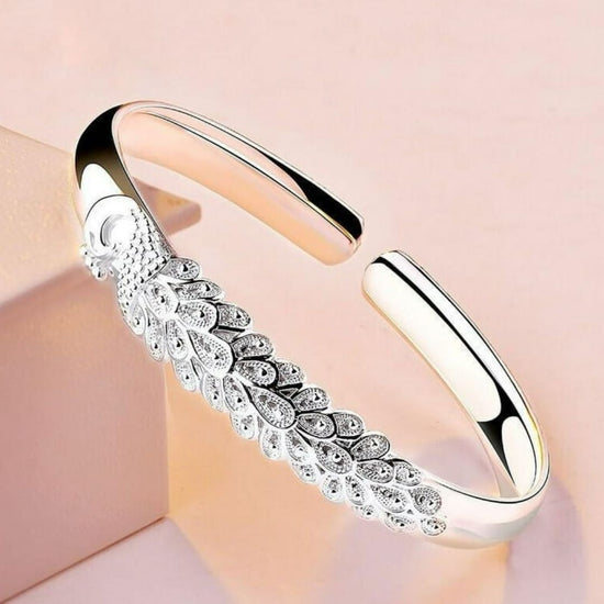 La Pulsera Elegancia Real es una pulsera abierta de plata con un intrincado dibujo de plumas de pavo real y pequeñas piedras preciosas, bellamente realzadas sobre un fondo rosa claro.