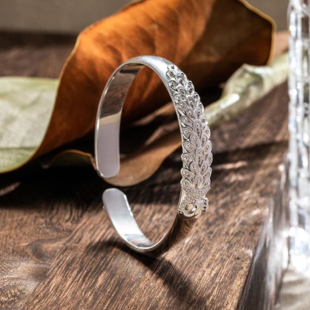 La Pulsera Elegancia Real, una pulsera de plata con un intrincado diseño inspirado en el pavo real, se alza sobre una superficie de madera junto a una hoja seca y un objeto de cristal al fondo.