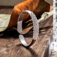 La Pulsera Elegancia Real, una pulsera de plata con un intrincado diseño inspirado en el pavo real, se alza sobre una superficie de madera junto a una hoja seca y un objeto de cristal al fondo.