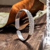 La Pulsera Elegancia Real, una pulsera de plata con un intrincado diseño inspirado en el pavo real, se alza sobre una superficie de madera junto a una hoja seca y un objeto de cristal al fondo.