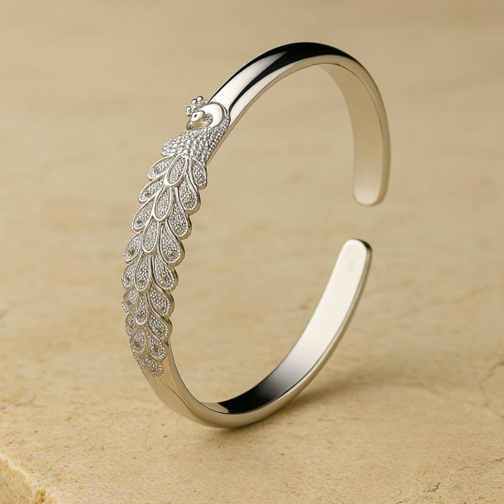 La Pulsera Elegancia Real es un brazalete de plata con un intrincado motivo de pavo real y plumas detalladas, lucido sobre una superficie de piedra beige.