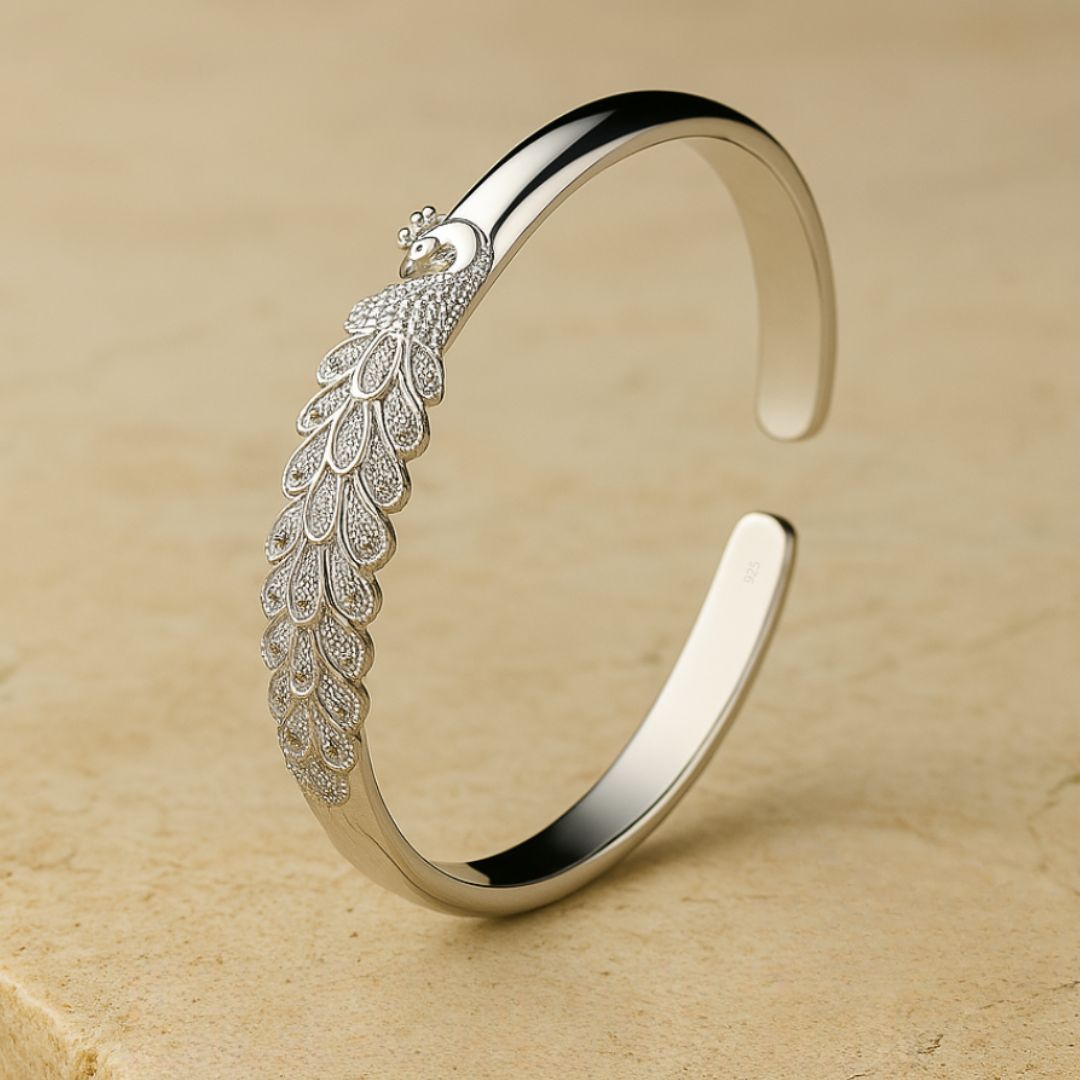 La Pulsera Elegancia Real es un brazalete de plata con un intrincado motivo de pavo real y plumas detalladas, lucido sobre una superficie de piedra beige.