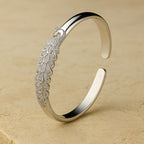 La Pulsera Elegancia Real es un brazalete de plata con un intrincado motivo de pavo real y plumas detalladas, lucido sobre una superficie de piedra beige.