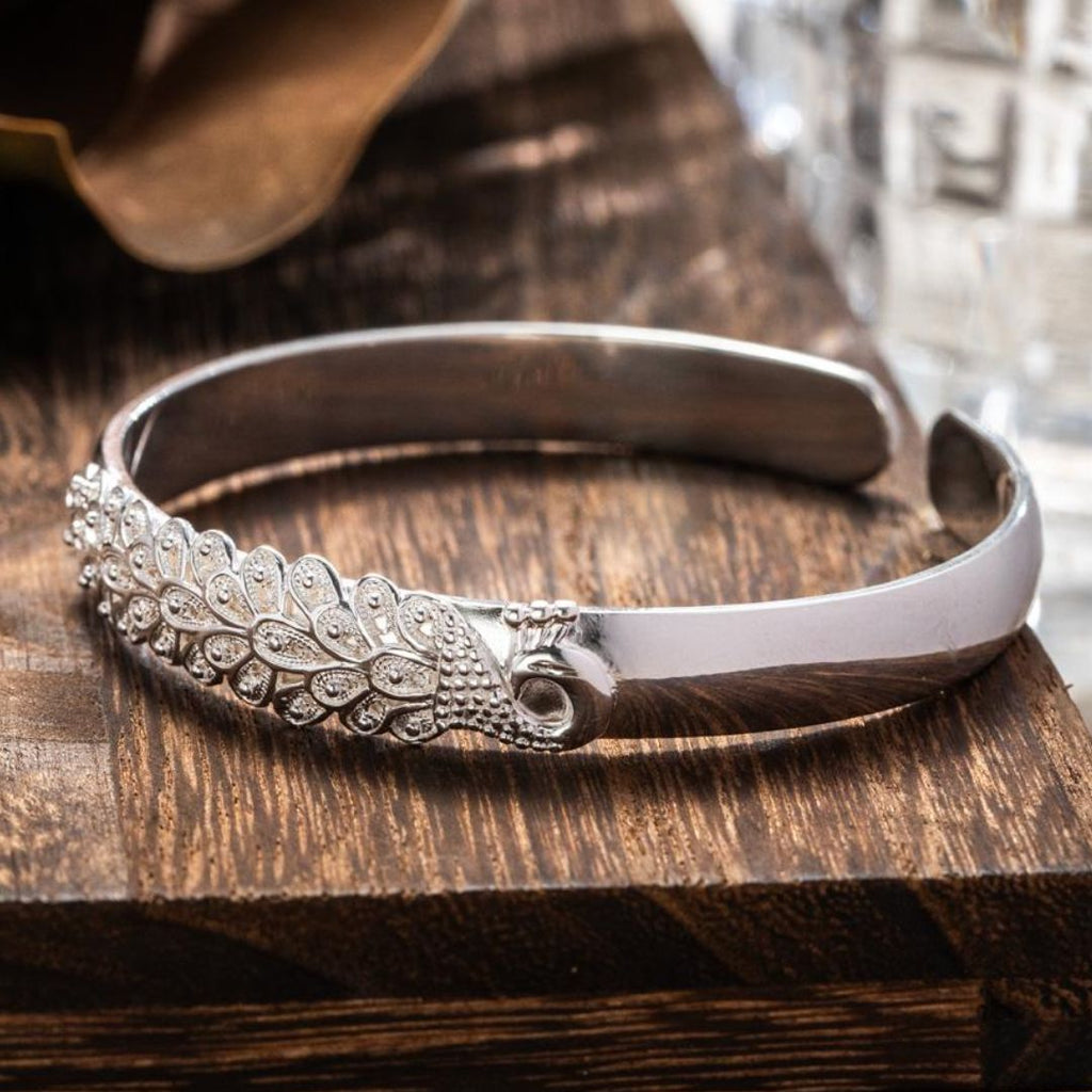 La Pulsera Elegancia Real es una pulsera de plata acero inoxidable con un diseño ornamentado en forma de hoja y pequeñas piedras incrustadas, elegantemente expuestas sobre una superficie de madera oscura.