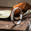 La Pulsera Elegancia Real, una pulsera de plata con detallados diseños florales y de hojas, se alza sobre una superficie de madera con hojas secas de fondo.