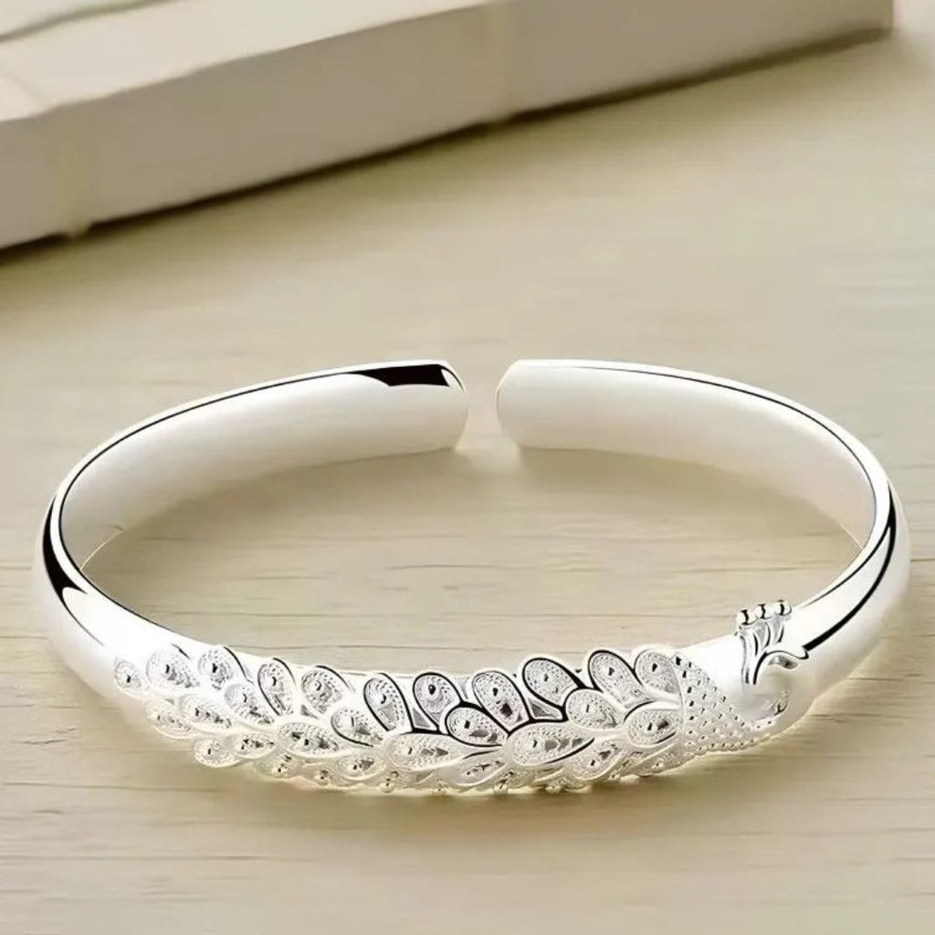 Pulsera Elegancia Real: una pulsera abierta de plata con un diseño grabado de pavo real y plumas, lucido sobre una superficie de madera clara.