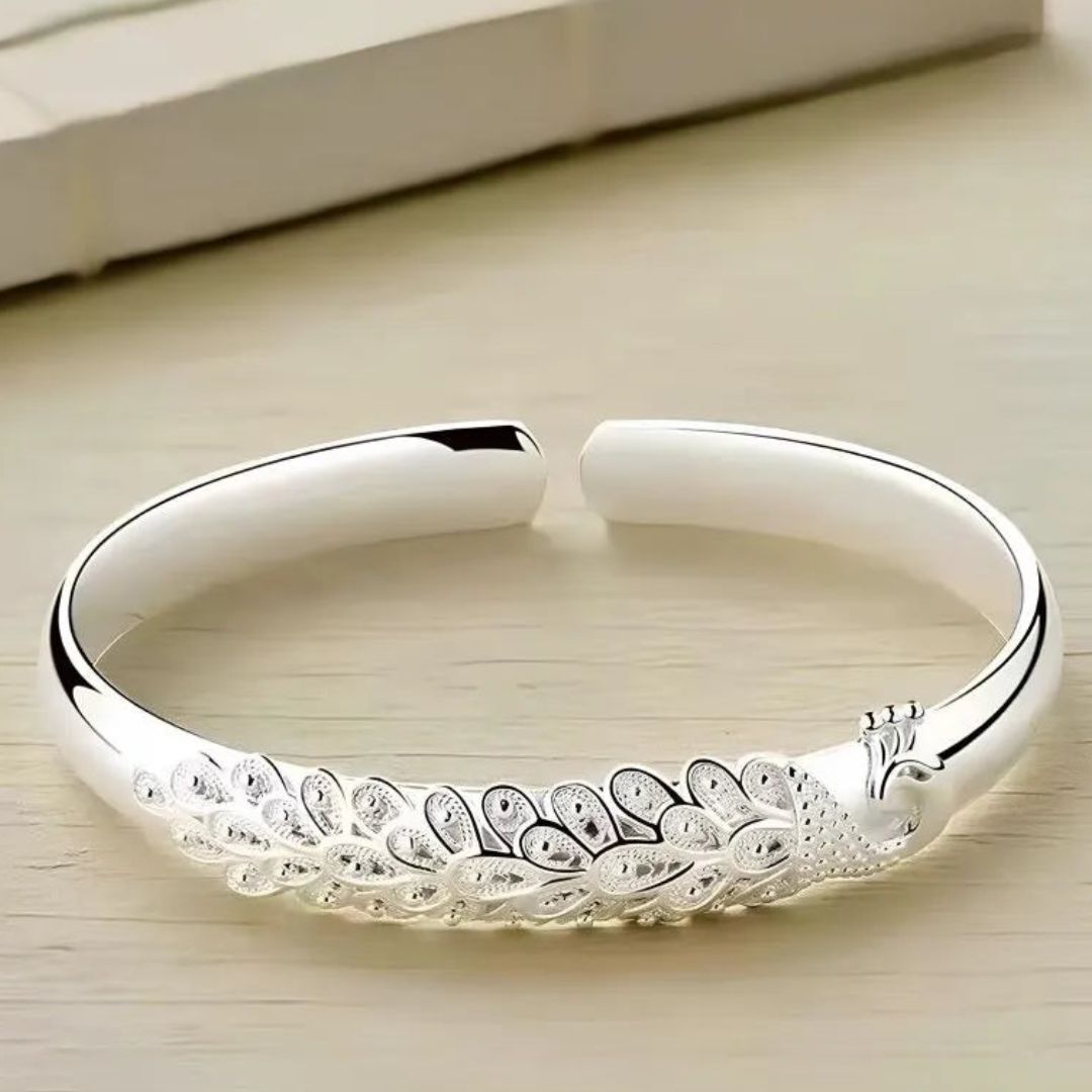 Pulsera Elegancia Real: una pulsera abierta de plata con un diseño grabado de pavo real y plumas, lucido sobre una superficie de madera clara.