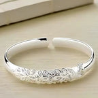 Pulsera Elegancia Real: una pulsera abierta de plata con un diseño grabado de pavo real y plumas, lucido sobre una superficie de madera clara.