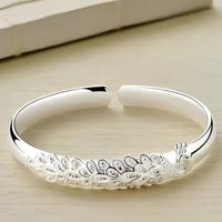 Pulsera Elegancia Real: una pulsera abierta de plata con un diseño grabado de pavo real y plumas, lucido sobre una superficie de madera clara.