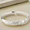 Pulsera Elegancia Real: una pulsera abierta de plata con un diseño grabado de pavo real y plumas, lucido sobre una superficie de madera clara.