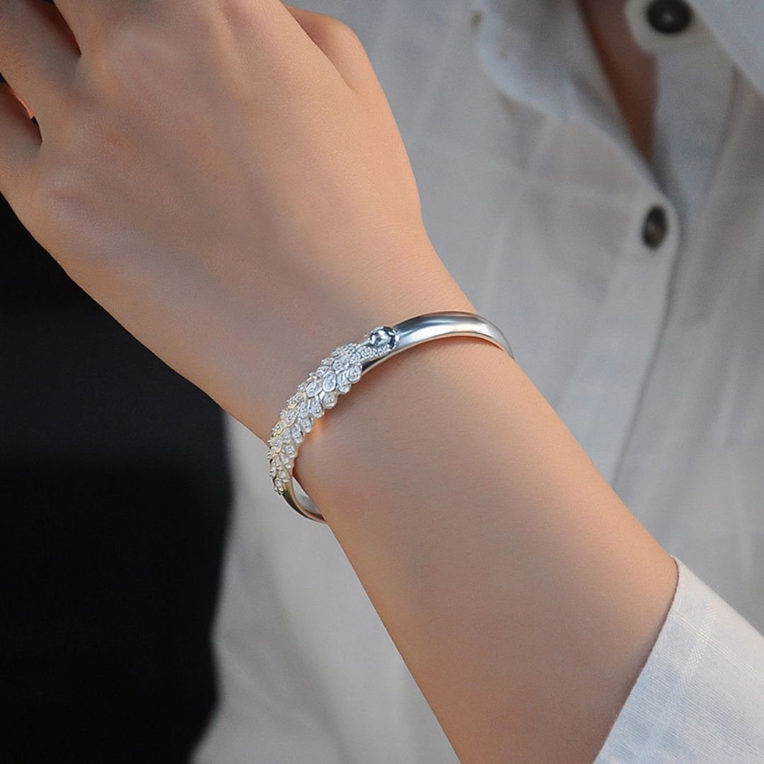 Una persona con camisa blanca luce en su muñeca la Pulsera Elegancia Real, un brazalete de acero inoxidable con un intrincado diseño inspirado en hojas y detalles brillantes.