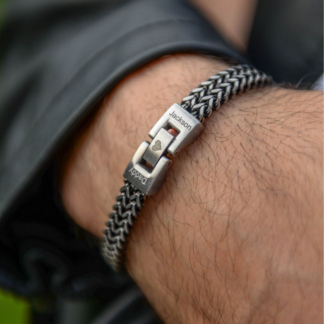 Un primer plano de una muñeca con la pulsera grabada - A mi amor, de cadena tejida plateada y broches personalizados con "Jackson", "Daddy" y un corazón. Un regalo masculino, ideal para quien lleva chaqueta de cuero negra.