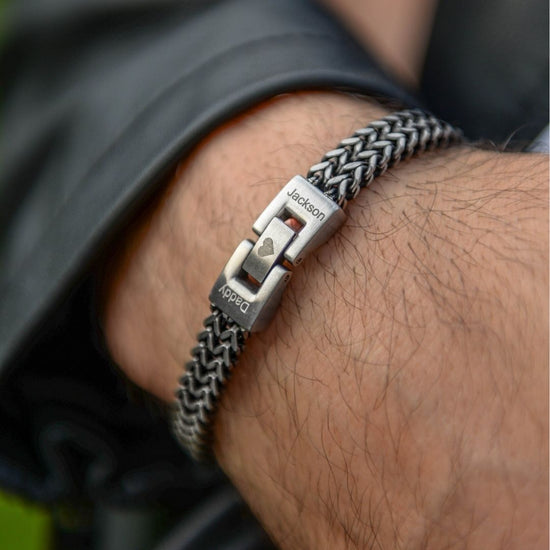 Un primer plano de una muñeca con la pulsera grabada - A mi amor, de cadena tejida plateada y broches personalizados con "Jackson", "Daddy" y un corazón. Un regalo masculino, ideal para quien lleva chaqueta de cuero negra.