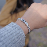 Primer plano de una muñeca con la Pulsera grabada - A mi amor, una pulsera de cadena de plata grabada con un mensaje especial. En el fondo difuminado se ve a alguien con una chaqueta marrón, lo que la convierte en un regalo ideal para hombre.