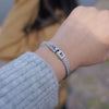 Primer plano de una muñeca con la Pulsera grabada - A mi amor, una pulsera de cadena de plata grabada con un mensaje especial. En el fondo difuminado se ve a alguien con una chaqueta marrón, lo que la convierte en un regalo ideal para hombre.