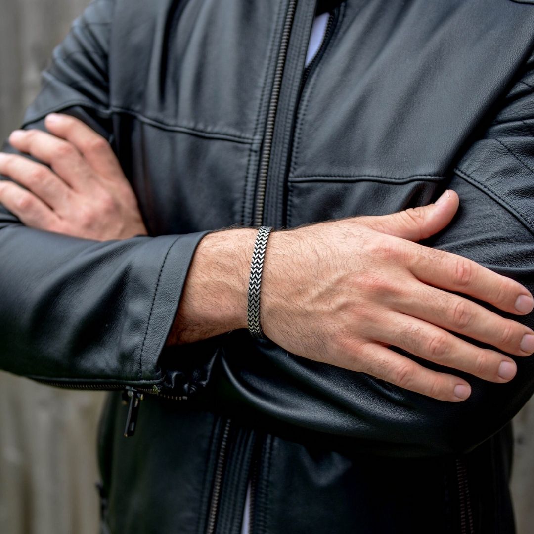 Una persona con una chaqueta de cuero negra cruza los brazos, mostrando la Pulsera grabada - A mi amor con un estampado chevron plateado. El fondo difuminado de paneles de madera lo convierte en un regalo ideal para hombre.