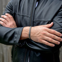 Una persona con una chaqueta de cuero negra cruza los brazos, mostrando la Pulsera grabada - A mi amor con un estampado chevron plateado. El fondo difuminado de paneles de madera lo convierte en un regalo ideal para hombre.