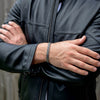 Una persona con una chaqueta de cuero negra cruza los brazos, mostrando la Pulsera grabada - A mi amor con un estampado chevron plateado. El fondo difuminado de paneles de madera lo convierte en un regalo ideal para hombre.