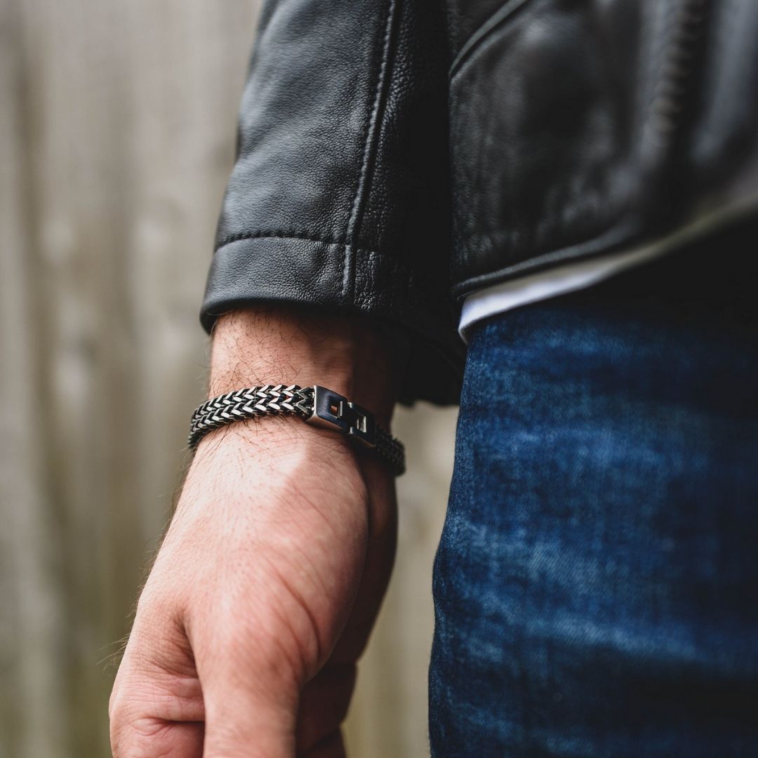 Primer plano de una mano y una muñeca que llevan la pulsera de cadena negra y plateada Pulsera grabada - A mi amor, combinada con una cazadora de cuero negra y unos vaqueros azules, un regalo ideal para hombre, sobre un fondo desenfocado.