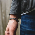 Primer plano de una mano y una muñeca que llevan la pulsera de cadena negra y plateada Pulsera grabada - A mi amor, combinada con una cazadora de cuero negra y unos vaqueros azules, un regalo ideal para hombre, sobre un fondo desenfocado.