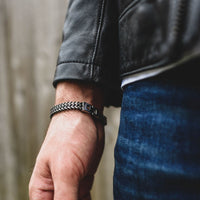 Primer plano de una mano y una muñeca que llevan la pulsera de cadena negra y plateada Pulsera grabada - A mi amor, combinada con una cazadora de cuero negra y unos vaqueros azules, un regalo ideal para hombre, sobre un fondo desenfocado.