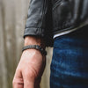 Primer plano de una mano y una muñeca que llevan la pulsera de cadena negra y plateada Pulsera grabada - A mi amor, combinada con una cazadora de cuero negra y unos vaqueros azules, un regalo ideal para hombre, sobre un fondo desenfocado.