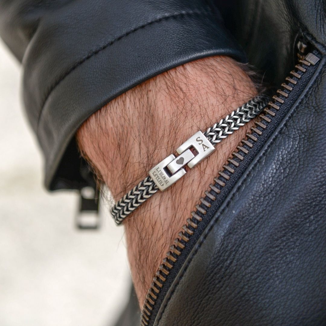 Primer plano de una persona con una chaqueta de cuero negro y la "Pulsera grabada - A mi amor", una pulsera de cadena de plata con un cierre rectangular grabado, en la muñeca.