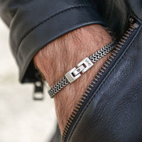 Primer plano de una persona con una chaqueta de cuero negro y la "Pulsera grabada - A mi amor", una pulsera de cadena de plata con un cierre rectangular grabado, en la muñeca.